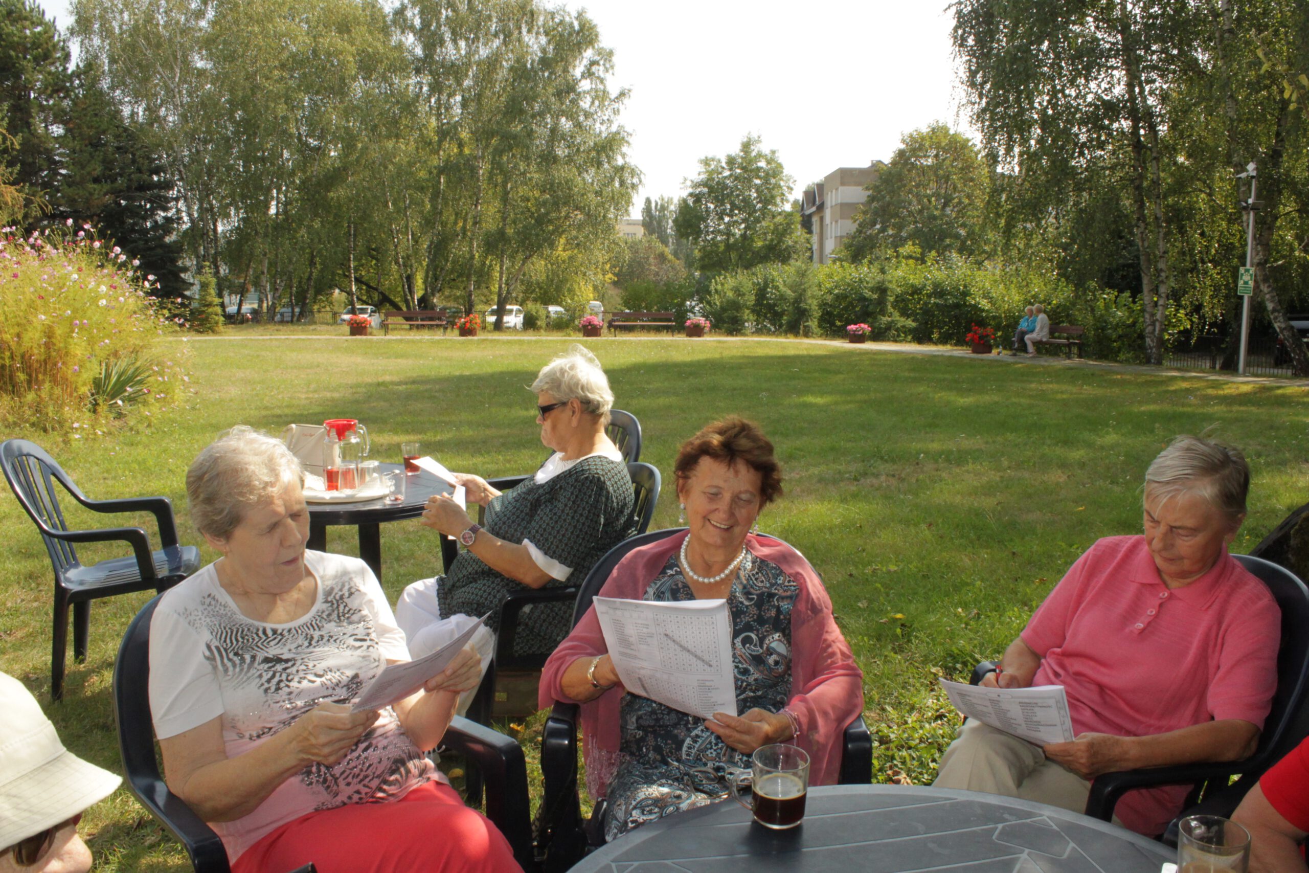 Seniorzy rozwiązujący krzyżówki, siedząc przy stolikach w ogrodzie Dziennego Domu.