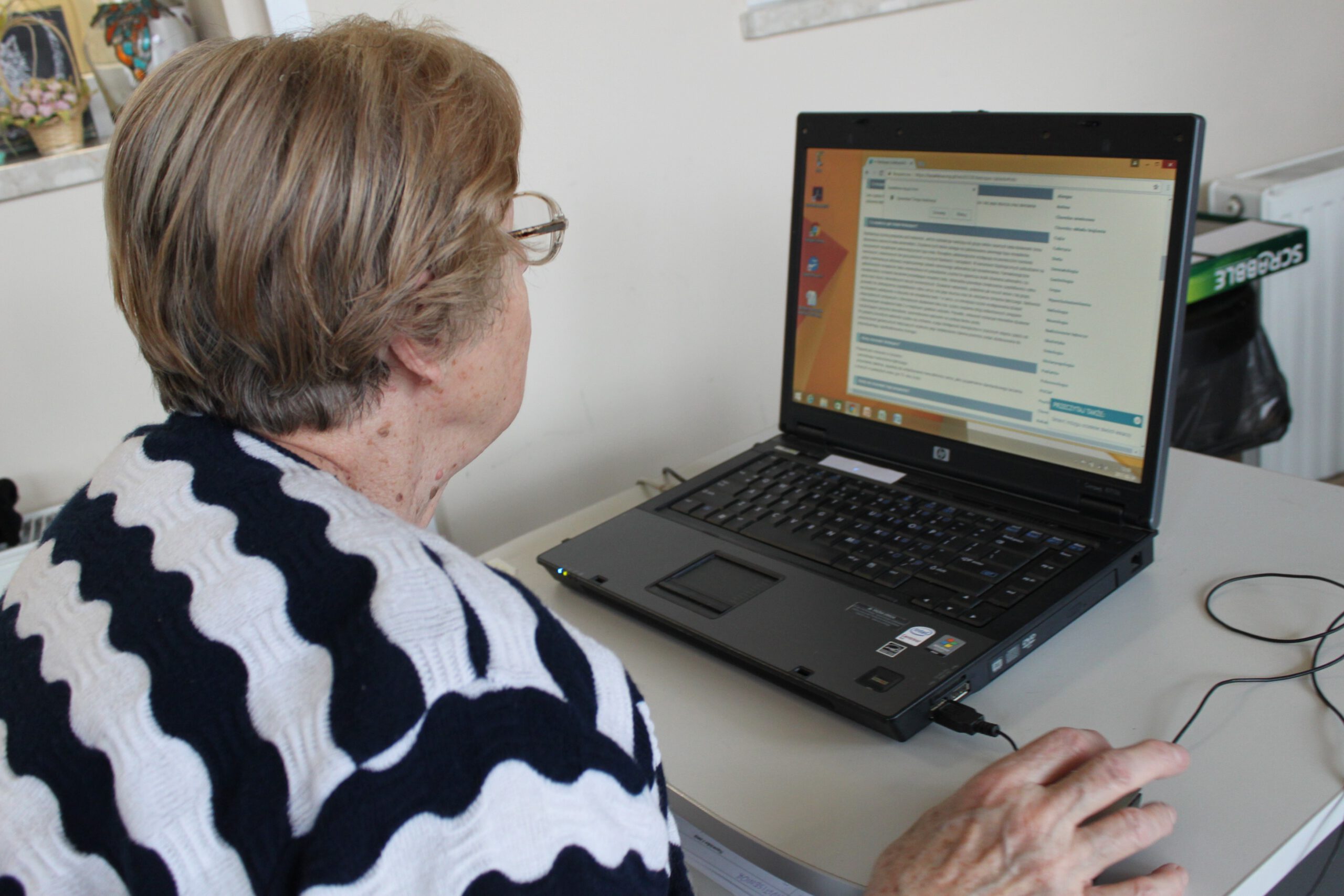 Seniorka Dziennego Domu siedzi przed laptopem i czyta artykuł w Internecie.